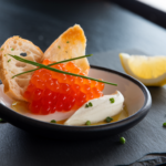 A beautifully plated salmon roe dish with crème fraîche, chives, and toasted bread on a black ceramic plate.